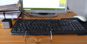Foto: Im Vordergrund eine schwarze Computertastatur auf einem Holztisch, halb darauf liegend eine Brille. Am oberen Bildrand abgeschnitten ein kleiner Computermonitor.